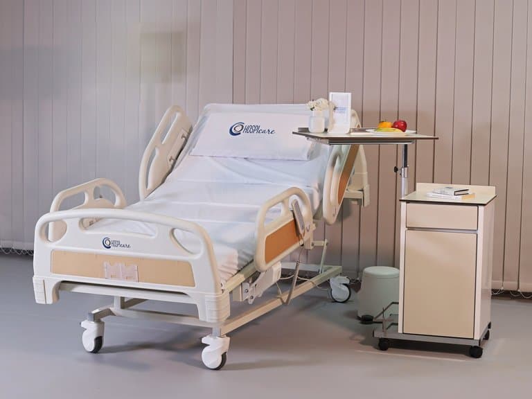 Multi-Functional PRIME care Bed in a furnished patient room with overbed table and bedside cabinet — MLP-01
